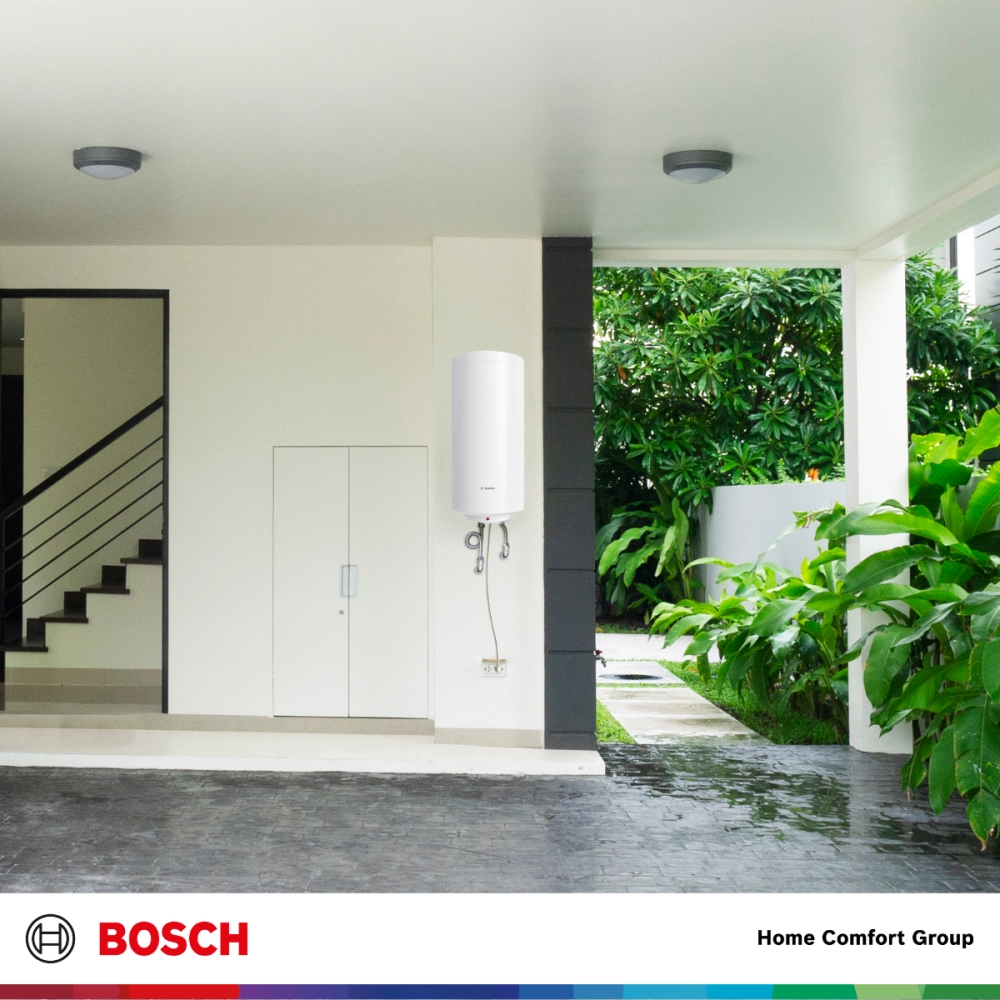 White cylindrical water heater mounted on an exterior wall next to a storage cabinet with a driveway and lush greenery in the background