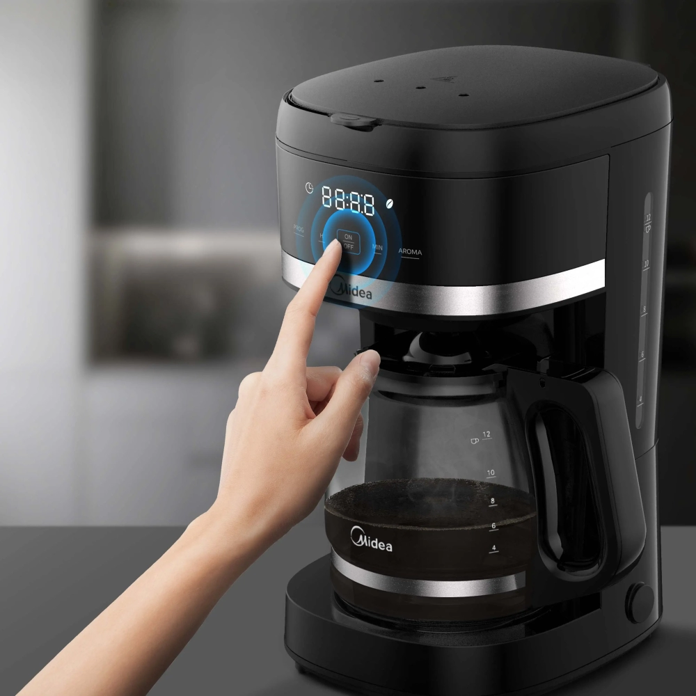 Person pressing a digital control on a black coffee maker to start brewing coffee at the front panel nearby a glass carafe with coffee inside